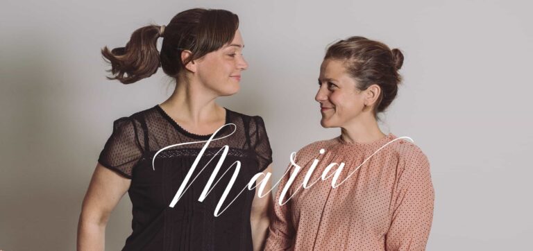 Zwei Frauen stehen vor einem schlichten Hintergrund, schauen sich an und lächeln. Die eine trägt ein schwarzes Top und hat die Haare zurückgebunden, die andere trägt ein rosa gemustertes Top und hat die Haare zu einem Dutt gebunden. Das Wort „Maria“ steht in Schreibschrift quer über das Bild – perfekt gestaltet von Grafikdesigner Aachen.