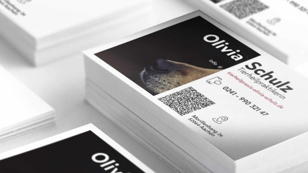 Auf einer weißen Fläche liegt ein Stapel Visitenkarten. Auf der obersten Karte steht in großen Buchstaben der Name „Olivia Schulz“ und ein Teilbild einer Hundeschnauze. Kontaktdaten und ein QR-Code sind ebenfalls sichtbar, zusammen mit einem Telefonsymbol und einer E-Mail-Adresse.
