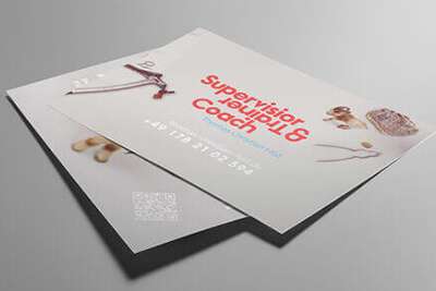 Auf einer grauen Fläche liegen zwei übereinanderliegende Flyer. Der obere Flyer, gestaltet von einem Grafikdesigner aus Aachen, trägt in roter Schrift den Text „Supervisor, Trainer u0026 Coach“, darunter einen QR-Code, eine Kontaktnummer und eine Website. Rund um die Flyer liegen verstreut Bastelutensilien und -materialien.