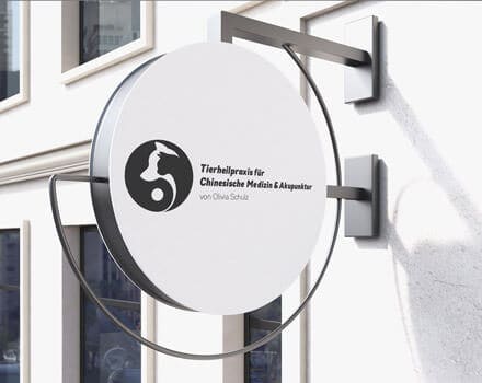 Das Bild zeigt ein rundes Schild für eine Tierarztpraxis mit Schwerpunkt Chinesische Medizin und Akupunktur von Olivia Schulz. Das von den Experten von Webdesign Aachen gestaltete Schild ist an einer Außenwand angebracht und zeigt ein schwarz-weißes Logo mit einem stilisierten Tier und deutschem Text.