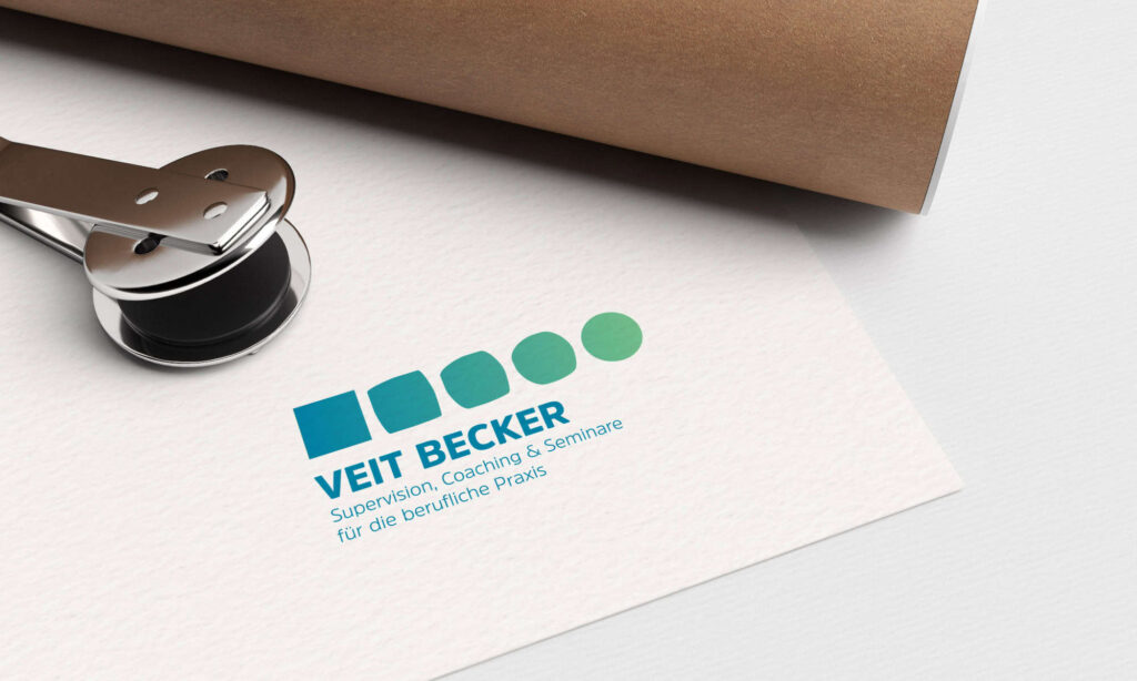 Ein Papier mit dem Logo „VEIT BECKER“ und dem Text „Supervision, Coaching u0026 Seminare für die berufliche Praxis“ in blauer Schrift. Das Logo besteht aus fünf Quadraten in Blautönen, die ins Grün übergehen. Eine Metallklammer presst das Papier gegen eine Rolle braunes Papier.