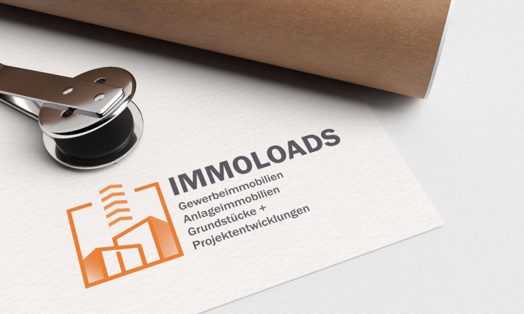 Ein Siegelstempel liegt neben einem Blatt Papier. Das Papier zeigt das IMMLOADS-Logo in orange-grauem Design mit einem Gebäude und geometrischen Formen. Der Text enthält deutsche Begriffe aus den Bereichen Immobilien und Projektentwicklung.