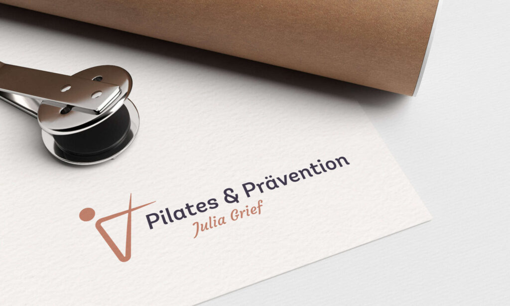 Eine Visitenkarte auf weißem Untergrund zeigt ein Logo mit einer stilisierten Figur und dem Text „Pilates u0026 Prävention Julia Grief“ in Marineblau und Pfirsich. Daneben liegen eine Metallklammer und ein brauner Ordner.